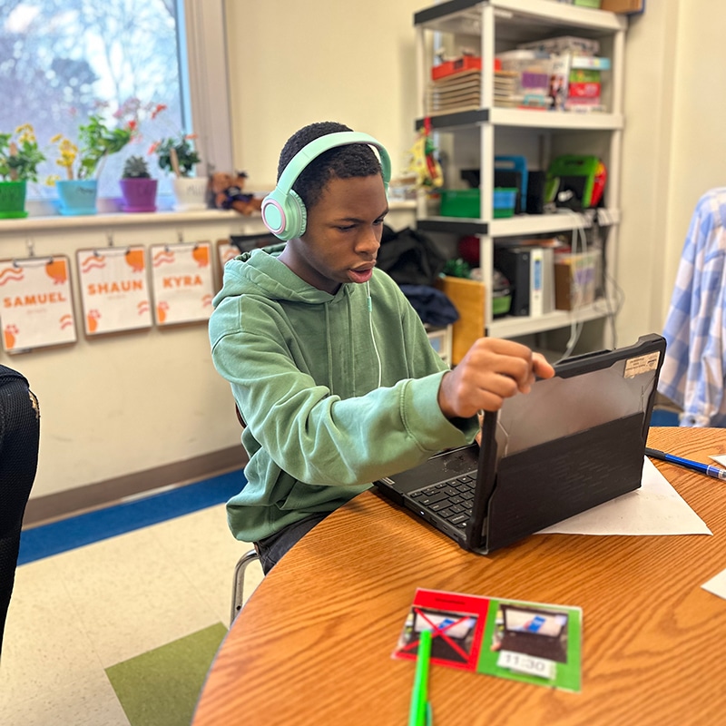 Student with green shirt and green headphones sitting at table using a laptop.